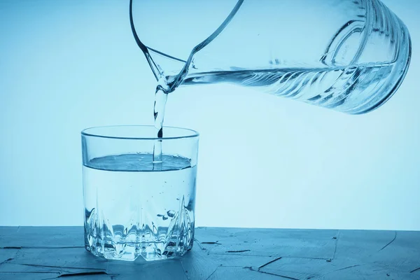 Pour water from a pitcher into a glass. Drinking water is poured from a jug into a glass on a blue background.