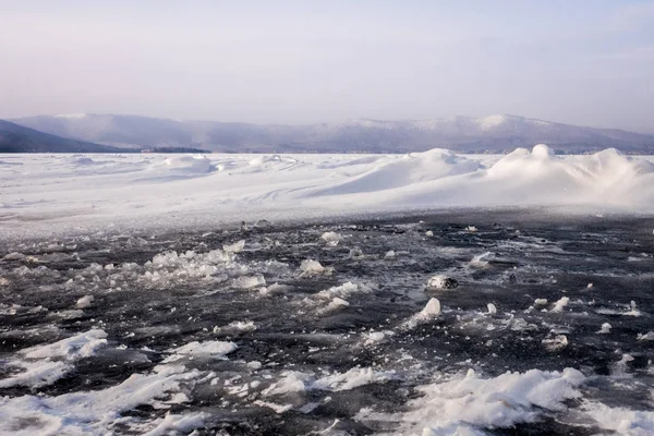 Natural winter lake ice breaking with clear sky background, selective focus. Frozen mountain lake with blue ice and cracks on the surface. Winter landscape with snowy hills under a blue sky.