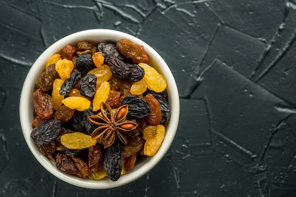 White ceramic bowl with Assortment of Raisins, yellow, blue, black, golden raisin on black background. Healthy snack. Vega Food. Food mix background, top view, copy space, banner.