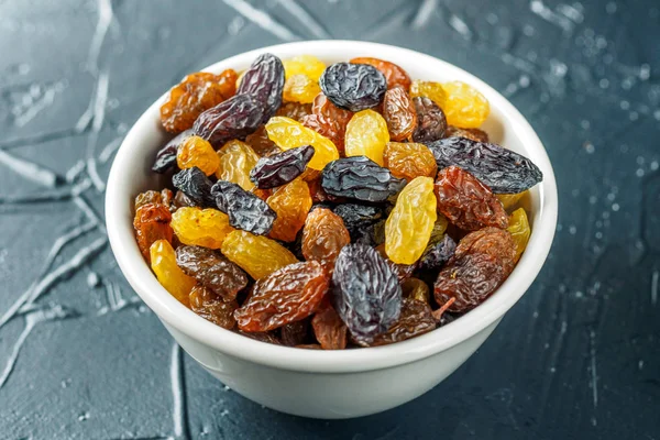 White ceramic bowl with Assortment of Raisins, yellow, blue, black, golden raisin on black background. Healthy snack. Vega Food. Food mix background, top view, copy space, banner.