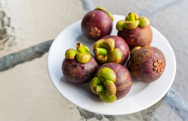 fresh mangosteen fruit pile lies on a white plate stands on a glass transparent table