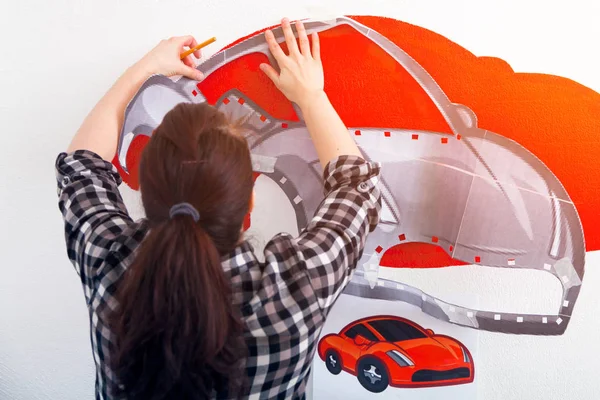 A close-up of a female artist draws a picture of a car and draws it in red on a white wall in the children\'s room