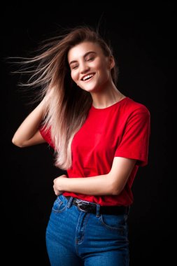 Kalça ve bakarak Studio fotoğraf makinesi üzerinde siyah arka plan üzerinde gülümseyen güzel genç kadın kırmızı t-shirt ve jaens ile poz eller