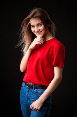 Kalça ve bakarak Studio fotoğraf makinesi üzerinde siyah arka plan üzerinde gülümseyen güzel genç kadın kırmızı t-shirt ve jaens ile poz eller