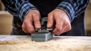 Close up of a experienced carpenter in work clothes and small buiness owner working in woodwork workshop, sharpening the tool with whetstone clipart
