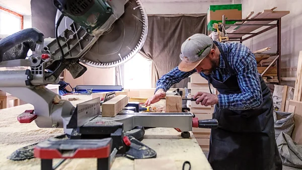 Experienced carpenter in work clothes and small buiness owner working in woodwork workshop, using a circular saw to cut through a wooden plank, on the table is a hammer and many tools