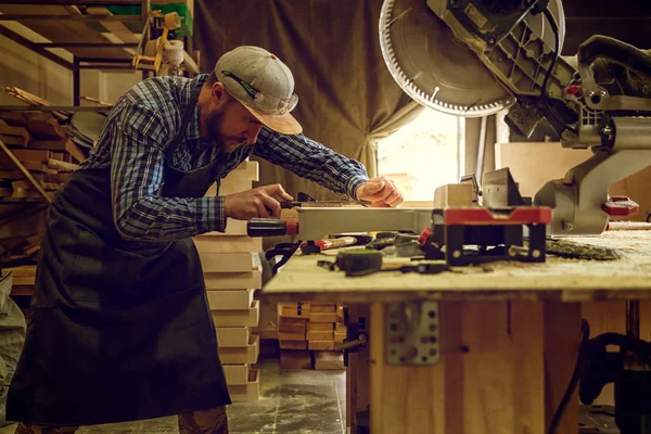 有经验的木匠在工作服装和小付多少营业税业主在木工车间工作 用圆锯切割木板 在桌子上是一个锤子和许多工具 — 图库照片