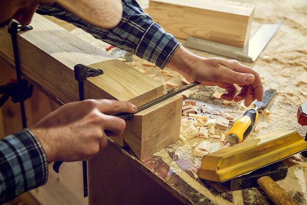 Close up experienced carpenter in work clothes and small buiness owner working in woodwork workshop,  using chisel for cutting out of wood  in workshop  on the table is a hammer and many tools 