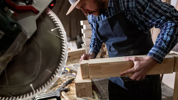 Carpenter in work clothes and small buiness owner working in woodwork workshop, using a circular saw to cut through a wooden, on the table is a hammer and many tools