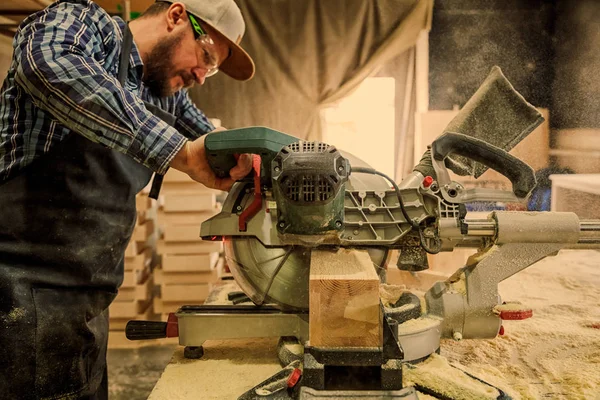 Experienced carpenter in work clothes and small buiness owner working in woodwork workshop, using a circular saw to cut through a wooden plank, on the table is a hammer and many tools