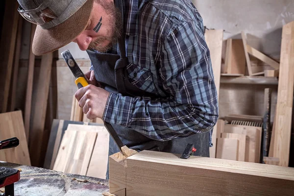 Close up experienced carpenter in work clothes and small buiness owner working in woodwork workshop,  using chisel for cutting out of wood  in workshop  on the table is a hammer and many tools