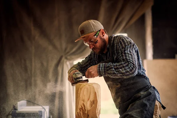 Experienced carpenter in work clothes and small buiness owner working in woodwork workshop, processes the board with an angle grinder on the table is a hammer and many tools