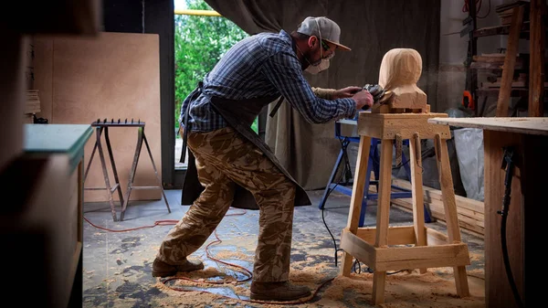 作業服で小さな事業所有者見た木造彫刻を男の頭をカットするワーク ショップは 多くの仕事のためのツール 家具のまわりでアングル グラインダーを使用 — ストック写真
