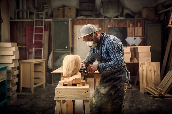 Close up of a carpenter, builder in work clothes saw to cut out sculpture from wooden a man\'s head, using an angle grinder  in the workshop, around a lot of tools,wooden,furniture for work