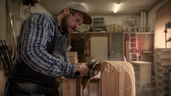 Carpenter in work clothes and small buiness owner working in woodwork workshop, processes the board with an angle grinder , on the table is a hammer and many tools