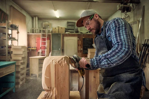 Carpenter in work clothes and small buiness owner working in woodwork workshop, processes the board with an angle grinder , on the table is a hammer and many tools