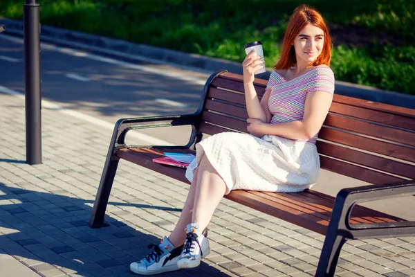 Bir dizüstü bilgisayar ve dizüstü etekli Beyaz Romantik, spor ayakkabı kahve içer, city Park, onun yanındaki bir yaz gününde bir bankta oturur bir pembe üst ve mavi spor ayakkabı Kızıl saçlı bir kadın olduğunu