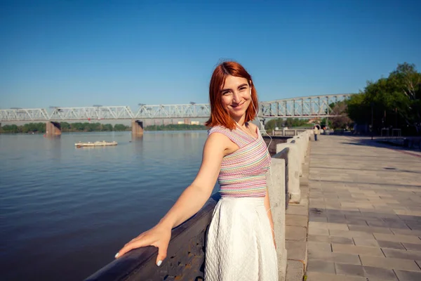 Kızıl saçlı kadın beyaz romantik etek, pembe bir üst telefonda müzik dinler ve bakar nehre şehir sahildeki bir yaz gününde