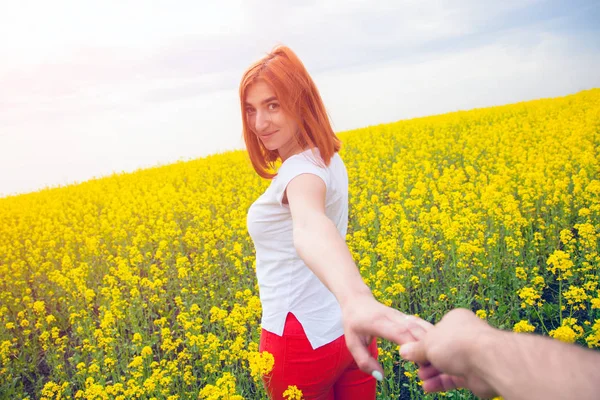 A young red-haired woman in a white T-shirt and red jeans smiles and leads the wet man\'s hand forward to the adventures of a huge field of yellow flowers on a warm summer day, follow me