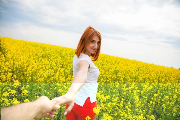 A young red-haired woman in a white T-shirt and red jeans smiles and leads the wet man\'s hand forward to the adventures of a huge field of yellow flowers on a warm summer day, follow me