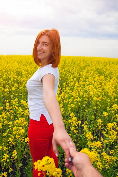A young red-haired woman in a white T-shirt and red jeans smiles and leads the wet man\'s hand forward to the adventures of a huge field of yellow flowers on a warm summer day, follow me