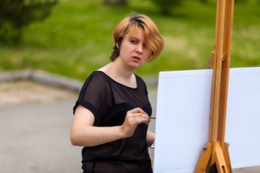 Bir genç sarışın kadın sanatçı olarak tuval ve bir şövale üzerinde siyah bir T-shirt ve jeans boyalar yağlı boya bir şehir parkı güneşli bir ile. Açık bir havada çizim