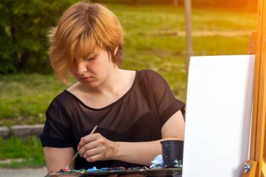 Bir genç sarışın kadın sanatçı olarak tuval ve bir şövale üzerinde siyah bir T-shirt ve jeans boyalar yağlı boya bir şehir parkı güneşli bir ile. Açık bir havada çizim