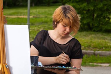 Bir genç sarışın kadın sanatçı olarak tuval ve bir şövale üzerinde siyah bir T-shirt ve jeans boyalar yağlı boya bir şehir parkı güneşli bir ile. Açık bir havada çizim