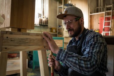 iş elbiseleri ve küçük buiness sahibi doğrama atölyesinde çalışan deneyimli marangoz bir tabure çivi çekiç için bir çekiç kullanmak