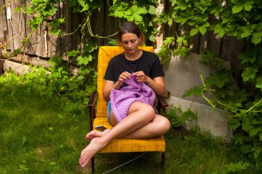 A young woman in a black T-shirt and jeans shorts knits a sweater from natural lilac woolen threads and sits on an armchair in the country on the background of a green lawn clipart