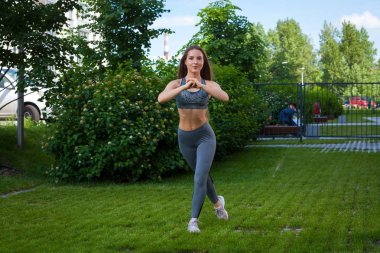 Bir esmer kadın eğitmen bir sportif kısa üst ve spor salonu tozluk lunges kalça önünde onu bir yaz gününde, elinde bir yeşil çim bir parkın pompalama için doğru tekniği gösterir
