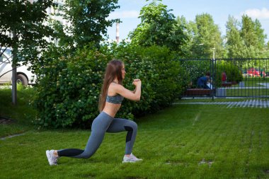 Bir esmer kadın eğitmen bir sportif kısa üst ve spor salonu tozluk kalça, tam bir ağız kavgası konumunu, seni elinde bir yeşil çim üzerinde bir parkta bir yaz gününde şişirmek için bacaklar lunging doğru tekniği gösterir