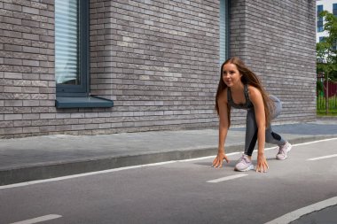 Bir sportif kısa üst ve spor salonu tozluk genç esmer kadın sporcumuz gülümsüyor ve bacaklarını bir yaz gününde bir modern kentsel avluda uzanıyor. 