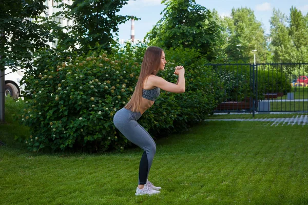 Doğru çömelme tekniği, başlangıç konumu, onun önünde yeşil çim üzerinde bir parkta bir yaz gününde eller esmer kadın bir sportif kısa üst ve spor salonu tozluk at arabasıyla gösterir bir genç