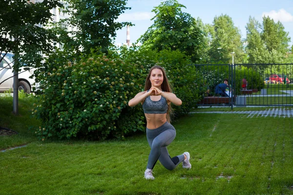 A dark-haired woman trainer in a sporty short top and gym leggings shows the correct technique of lunging the legs to inflate the buttocks, the position of a full squat, hands in front on a summer day in a park on a green lawn