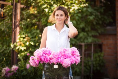 Beyaz bir gömlek genç bir esmer kadın bir bisiklet ile bir metal sepet peonies büyük bir buket ile yaz günü yeşil bir parkta duruyor