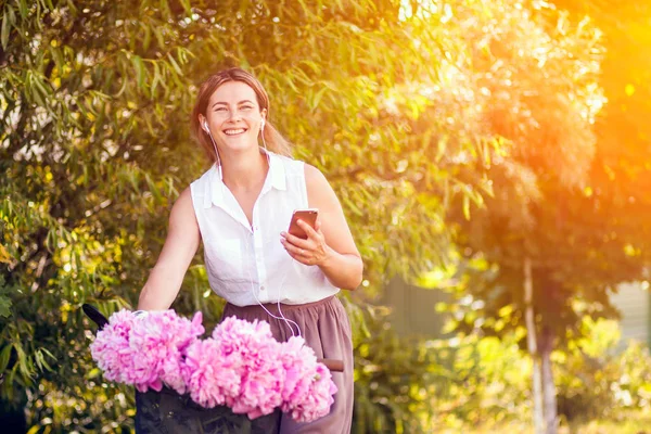 Young dark-haired woman in a white shirt in a beige skirt listens to music on the phone and stands with a bicycle with a metal basket with a big bouquet of peonies summer day in the park