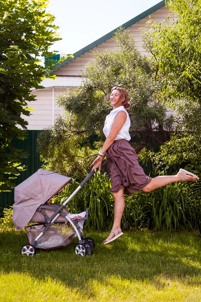 ベージュ スカートに白シャツに黒髪女性は小さな赤ん坊をベビーカーで歩くと明るい夏の日の公園で楽しみをジャンプ — ストック写真