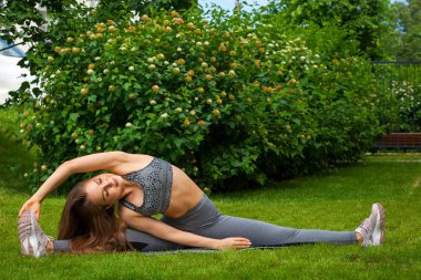 Esmer kadın at arabasıyla bir sportif kısa üst ve spor salonu tozluk yoga için halının üstünde geniş bir sicim yapar; Eller yukarı doğru yoga mat yetiştirilir, eller yukarı, yeşil çim üzerinde bir parkta bir yaz gününde yetiştirilir