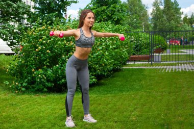 Bir sportif kısa üst ve spor salonu tozluk esmer kadın arabasıyla bir salıncak dumbbells ile iki tarafın elinde bir yaz gününde yeşil çim üzerinde bir parkta yapar