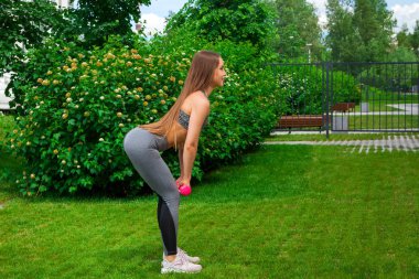 Sportif kısa üst ve spor salonu tozluk at esmer kadın arabasıyla bir deadlift dumbbells ile bir yaz gününde yeşil çim üzerinde bir parkta yapar