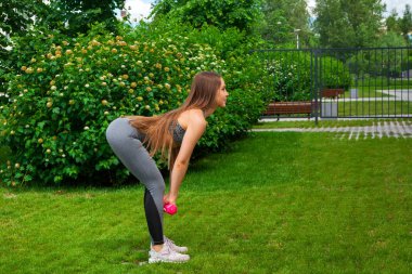 Sportif kısa üst ve spor salonu tozluk at esmer kadın arabasıyla bir deadlift dumbbells ile bir yaz gününde yeşil çim üzerinde bir parkta yapar