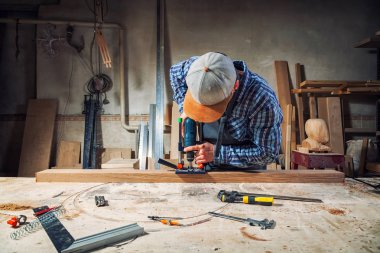 İş elbiseleri ve küçük buiness sahibi deneyimli marangoz araçları bir sürü ışık atölye yan görünümünde, arka planda bir modern el matkap ahşap bir gemide oyma