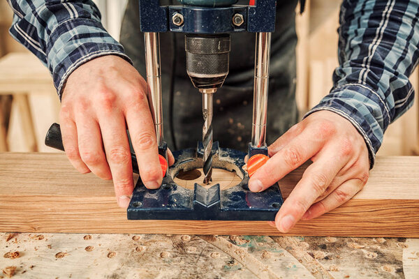 Крупным планом опытного плотника в рабочей одежде и владельца малого бизнеса является вырезание деревянной доски на современной ручной дрели в светлой мастерской вид сбоку, на заднем плане много инструментов
