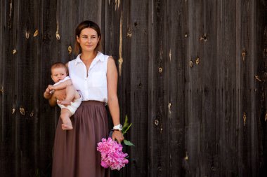 Güzel esmer kadın beyaz gömlek ve bej pembe peonies ahşap duvarlar arka plan üzerinde büyük bir buket ile gülümseyen, poz ve onun elinde tutan bebek etek