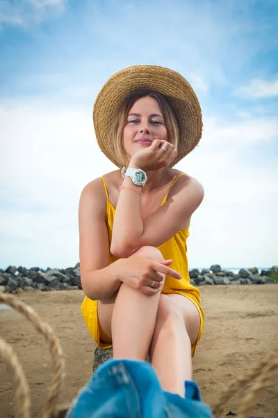 軽いカジュアルな服装と休憩とポーズを夏の日に青い空 青い空と美しい海の近くの砂浜のビーチの岩に麦わら帽子の若い美しい女性 — ストック写真