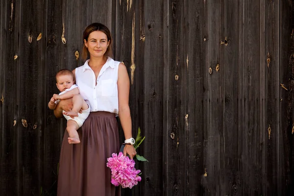 Güzel esmer kadın beyaz gömlek ve bej pembe peonies ahşap duvarlar arka plan üzerinde büyük bir buket ile gülümseyen, poz ve onun elinde tutan bebek etek