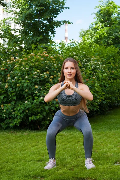 一个黑发女子教练在运动的短顶和健身房紧身裤显示正确的蹲技术 在一个完整的蹲下的位置 在一个夏天的一天 在公园的绿色草坪上的手 — 图库照片