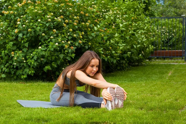 A dark-haired woman coach in a sporty short top and gym leggings does stretching the legs on the yoga mat,   on a summer day in a park on a green lawn