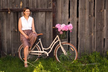 Bir bej beyaz bir gömlek genç bir esmer kadın etek gülümsüyor ve ahşap bir çit karşı bir hasır sepet pembe peonies güzel bir buket ile bej bir retro Bisiklet oturur 